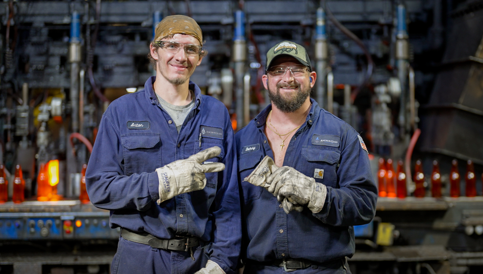 Two Anchor Glass operators in navy uniforms and work gloves smiling on the plant floor beside a hot end machine